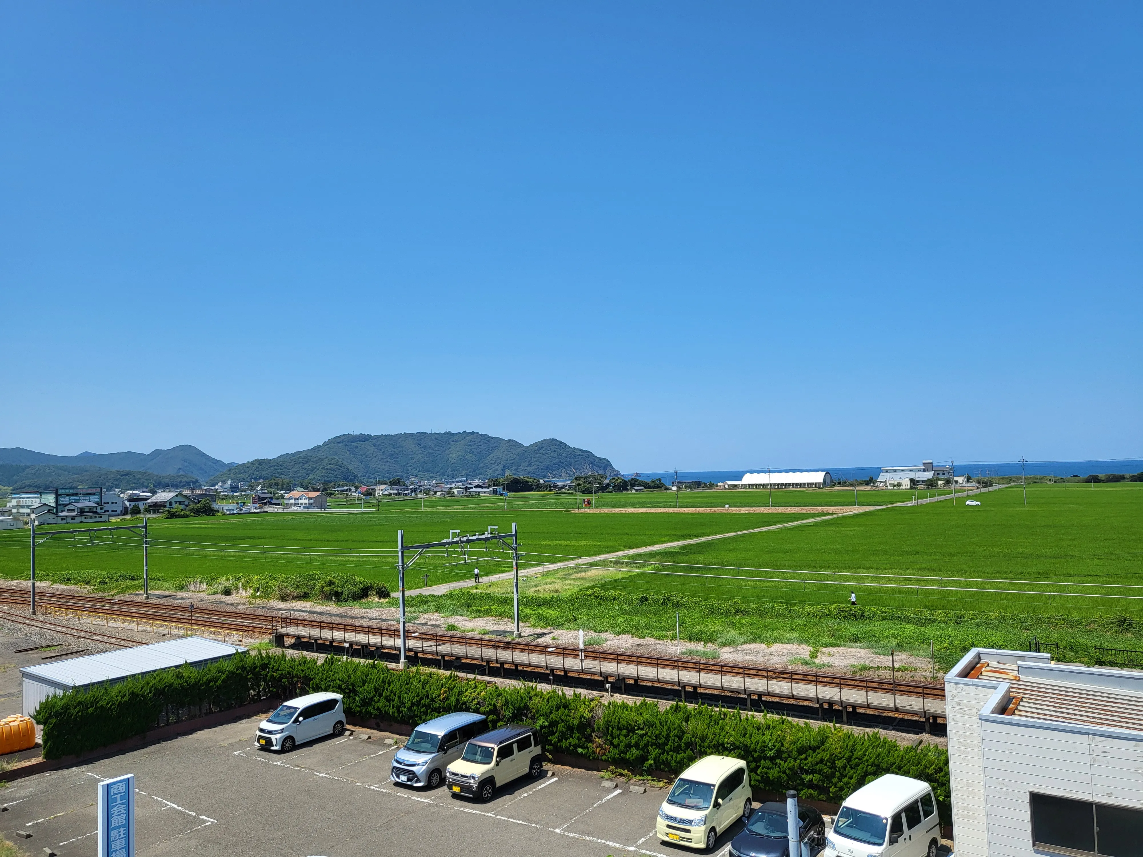 Green rice paddies stretching to the sea, with a small rural train line and hills in the distance